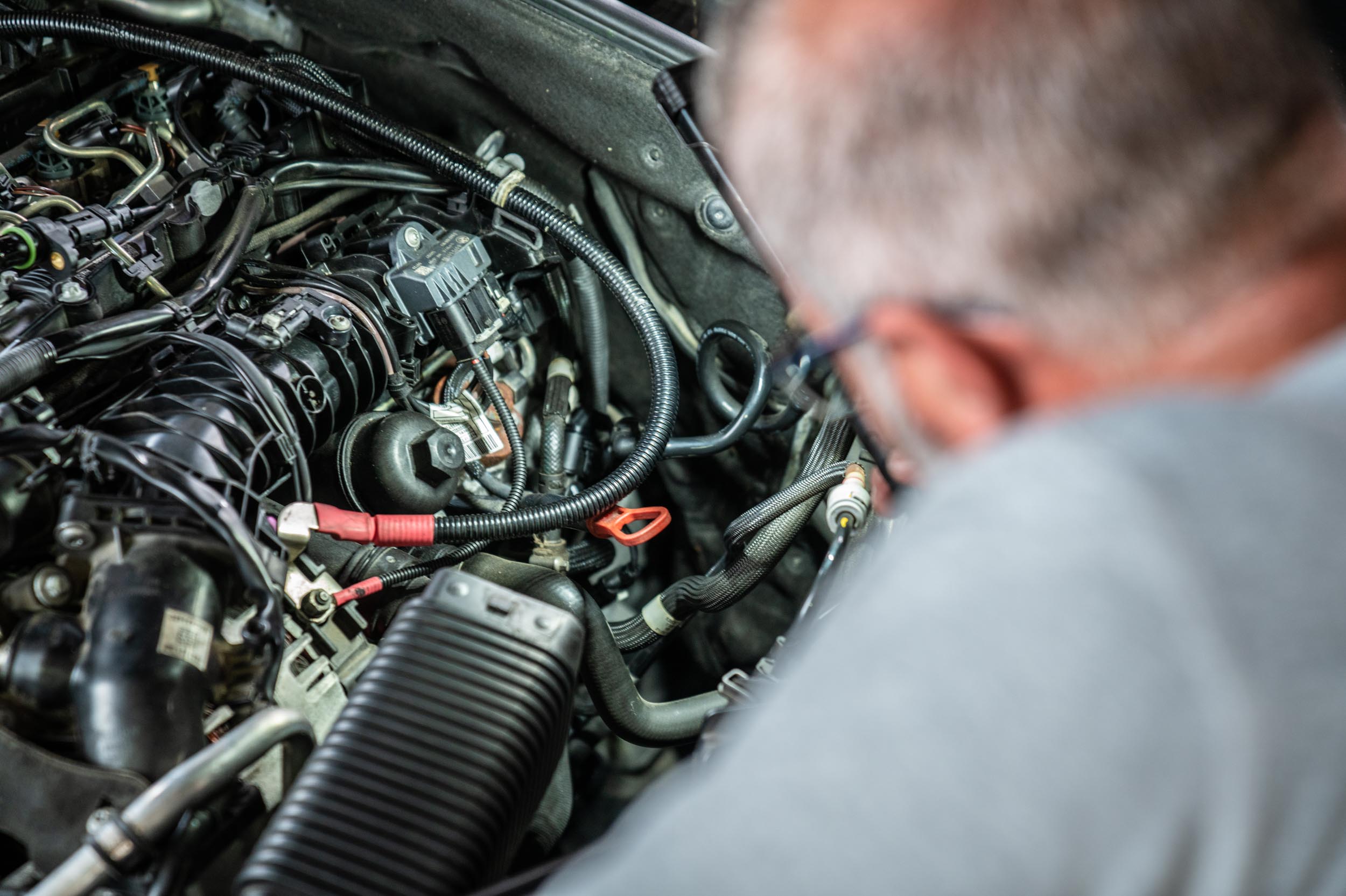 Nahaufnahme eines geöffneten Motorraums, an dem ein Mechaniker arbeitet. Zu sehen sind Kabel, Schläuche und Motorkomponenten, während die Person sich über den Motor beugt.