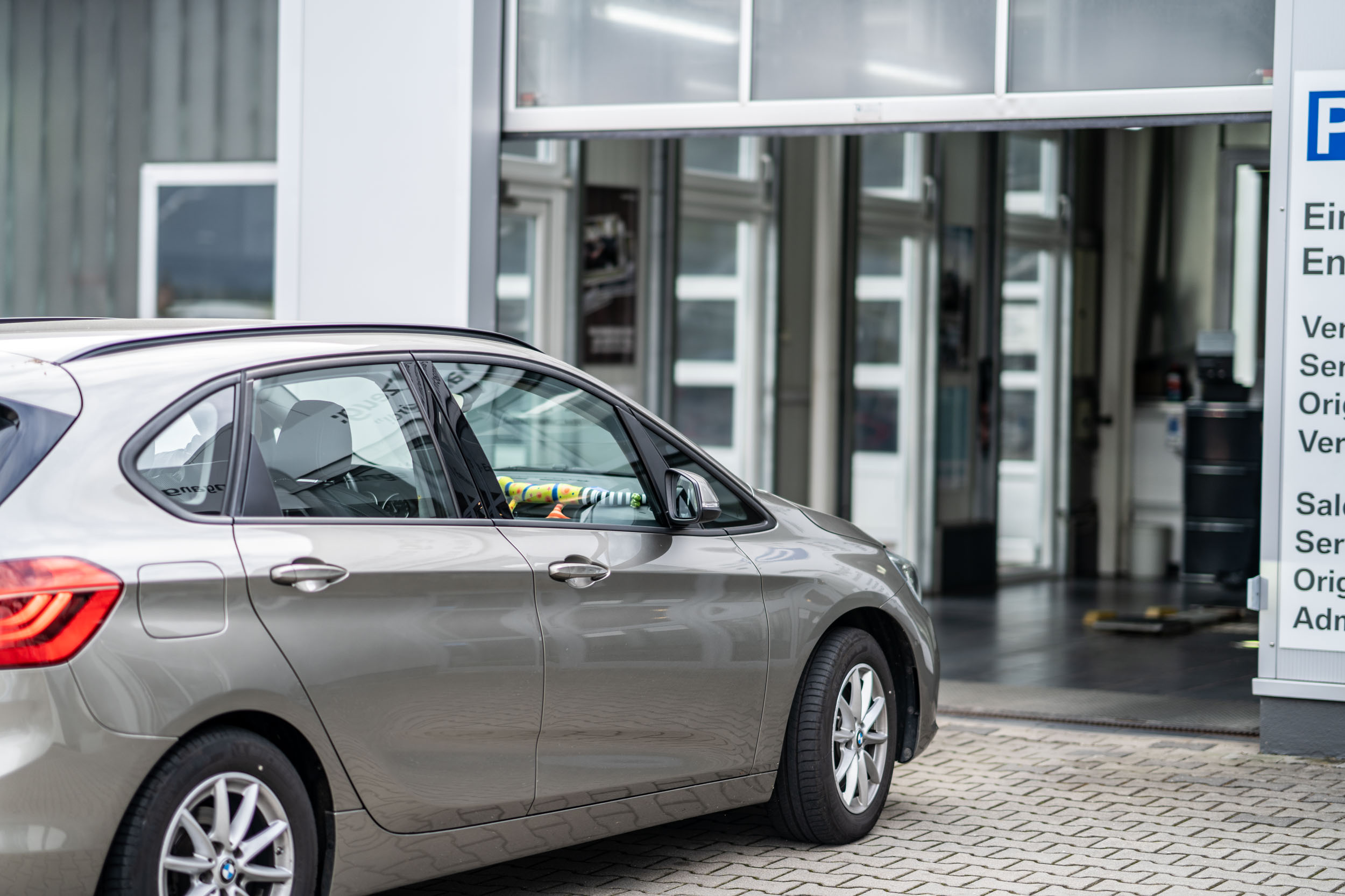 Ein grauer BMW steht vor der geöffneten Einfahrt einer Werkstatt. Im Hintergrund sind Glas- und Rolltore sowie ein Hinweisschild für den Servicebereich zu sehen.